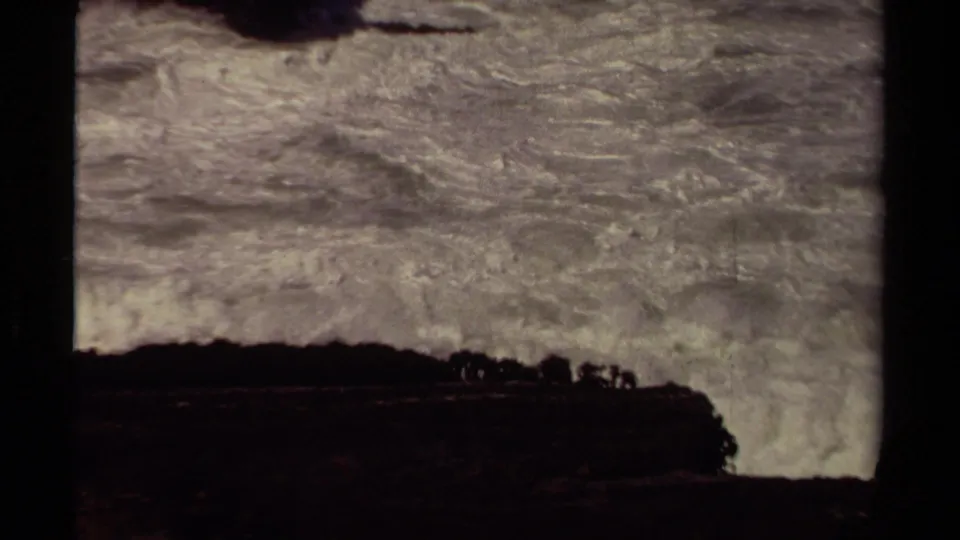 1980 coastal home-movie frame with turbulent white surf below the edge of a dark rock formation.