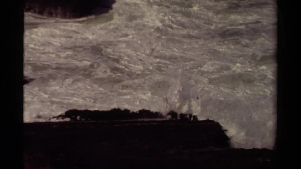 Mendocino 1980 8mm frame showing rolling white surf beneath the shadowed edge of a rock formation.