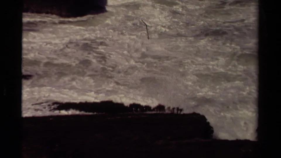 1980 Mendocino home-movie frame showing stormy Pacific surf breaking against a dark headland with tiny cliff-edge silhouettes.