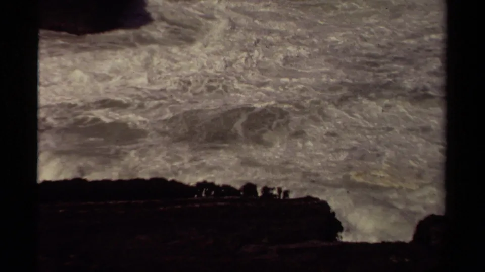 1980 Mendocino 8mm frame showing foamy storm waves surging below a dark coastal cliff with small figures watching.