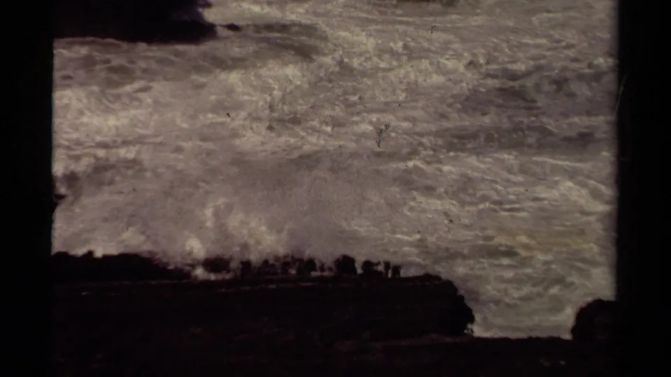 1980 Mendocino coastal home-movie frame with rough Pacific surf, a shadowed headland, and distant people along the cliff edge.