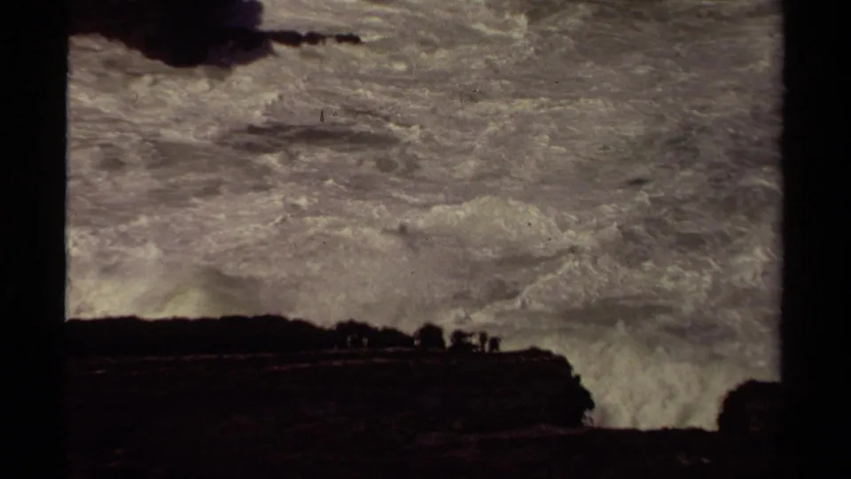 1980 8mm Mendocino coastal view with surf crashing into dark cliffs beneath a cloudy, faded sky.