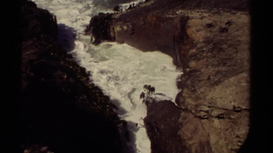 1980 Mendocino home-movie frame with foamy water moving through a rugged rock channel under uneven natural light.