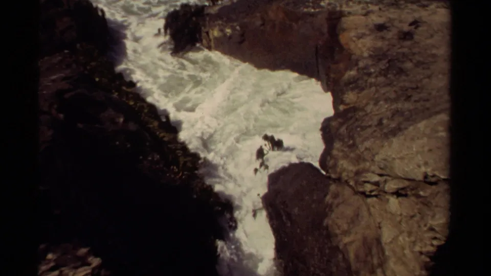 1980 8mm view of Mendocino surf surging between dark rocks, with small distant figures visible on the shoreline.