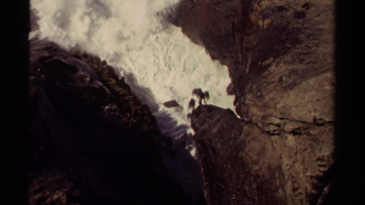 Waves break hard against rocks with distant figures near the cliff edge.