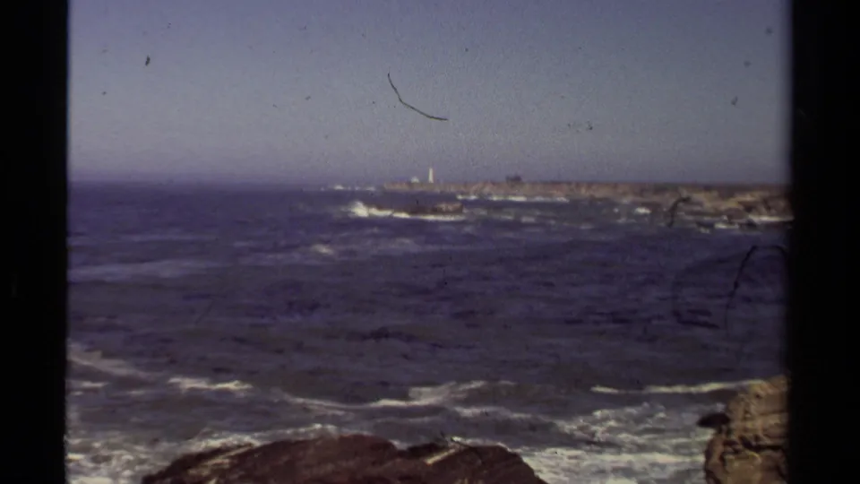 Mendocino coastal view with ocean surf, rocky shore, and a distant lighthouse-like structure on an island.