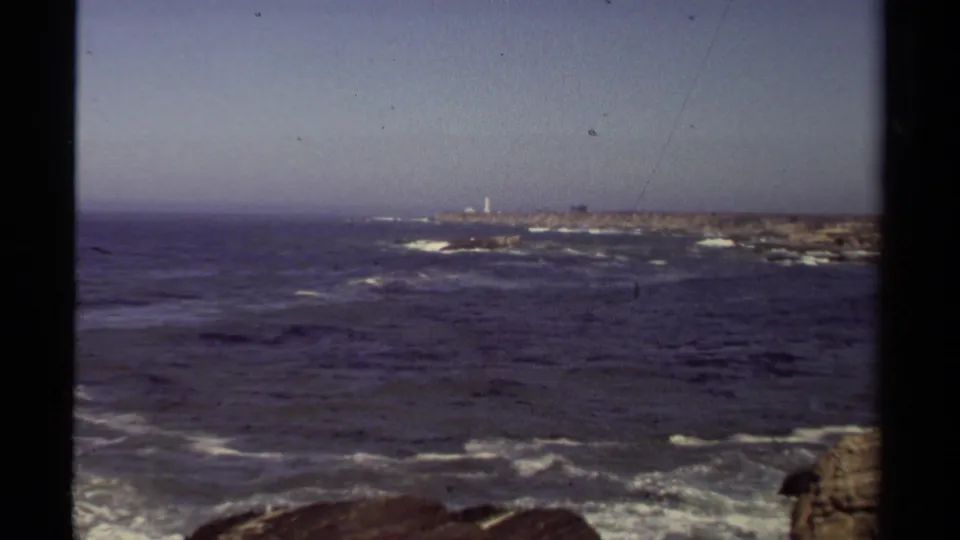 1980 home-movie view of the Mendocino coast with dark ocean, white surf, rocky shore, and a small distant lighthouse-like structure.