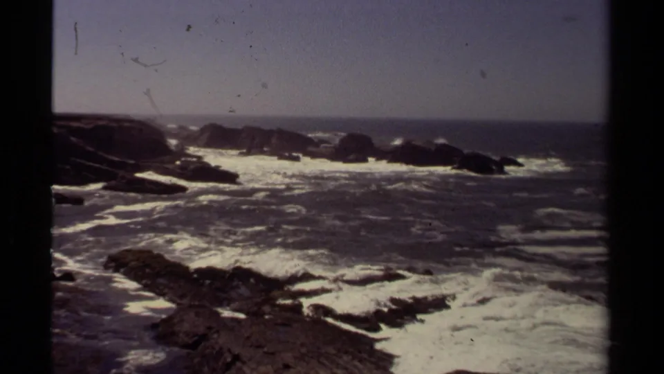 Vintage 8mm view of Mendocino surf with rough water, white foam, foreground rocks, and a dark rocky outcrop offshore.