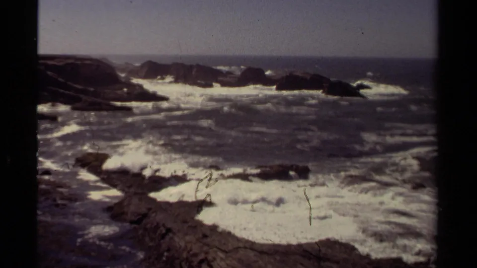 1980 Mendocino 8mm frame showing Pacific surf crashing around dark rocky outcrops from a high shoreline viewpoint.