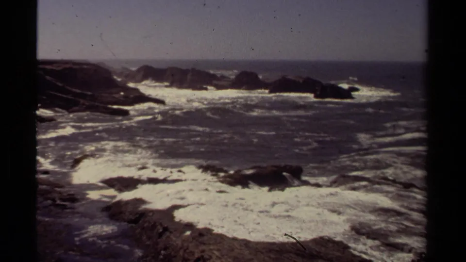 1980 Mendocino coastal frame with white Pacific surf washing over jagged rocks beneath a clear, pale sky.
