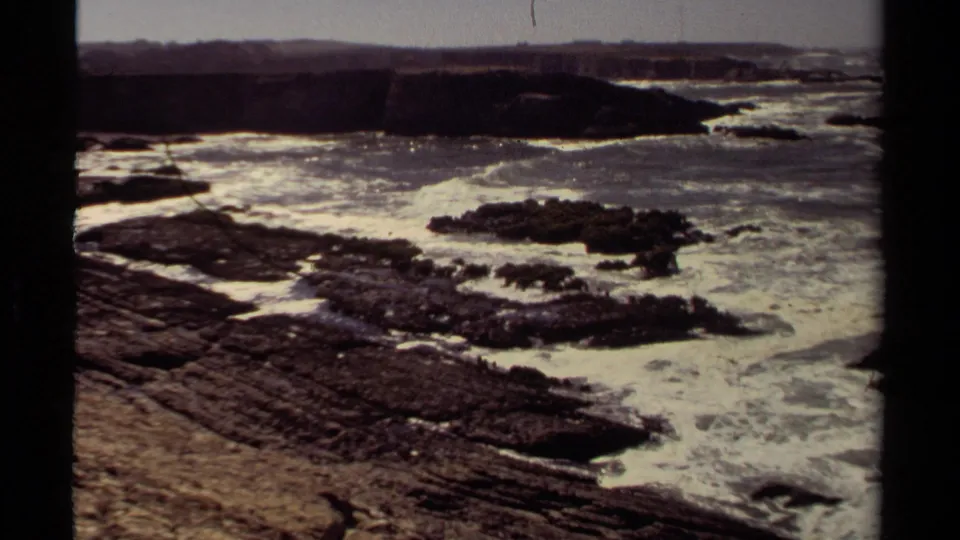1980 Mendocino coastal frame showing foamy Pacific waves around dark rock shelves and a low distant shoreline.