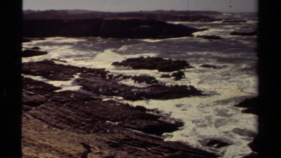 1980 8mm view of waves foaming over dark layered rocks on the Mendocino, California coast.