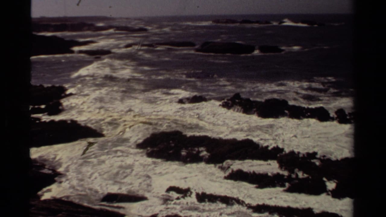 Dark ocean waves breaking around jagged rocks beneath a muted horizon.