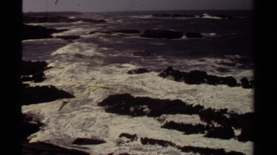 1980 Mendocino 8mm frame showing rough surf and white foam moving around dark coastal rocks under a dim sky.