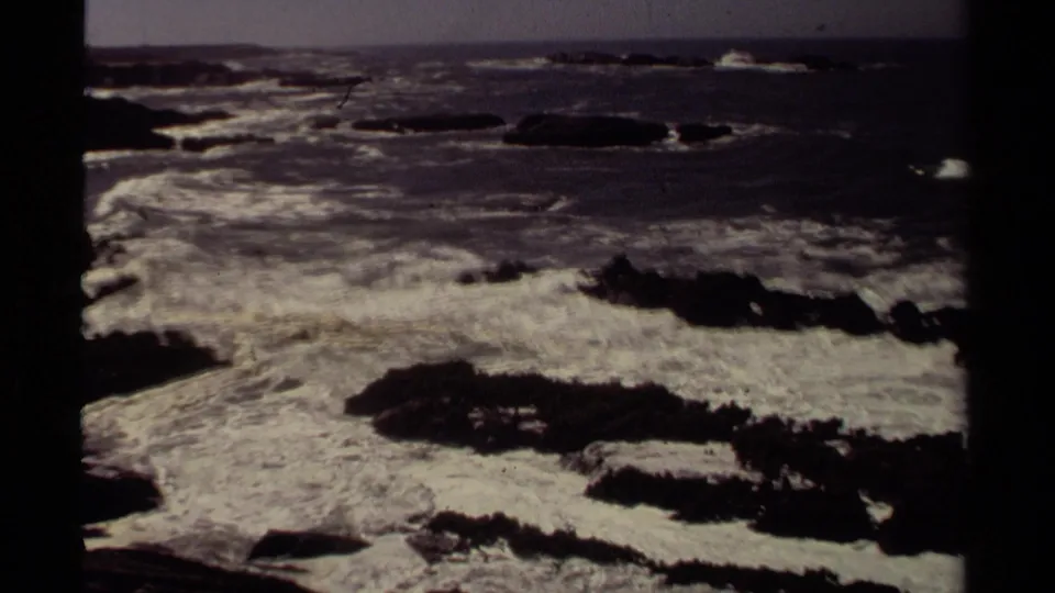 1980 Mendocino coastal frame with white surf churning over black rocks and darker ocean water in the distance.