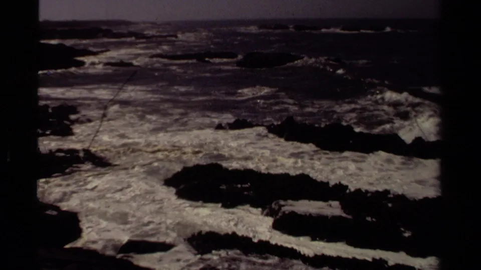 1980 Mendocino frame showing dark exposed rocks and pale surf under a subdued coastal horizon.