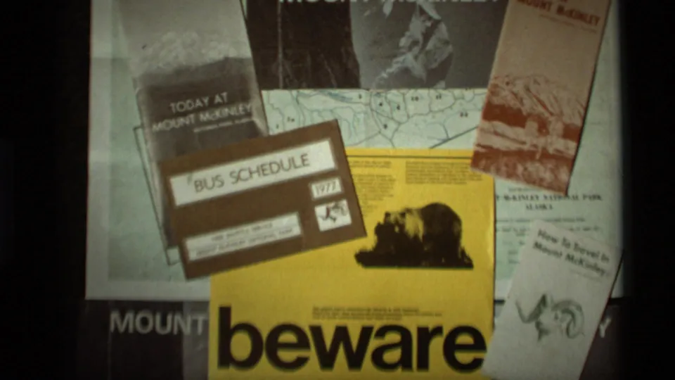 Visitor-shot 8mm frame showing a 1977 bus schedule placed over Mount McKinley travel papers and a bear safety warning sign.
