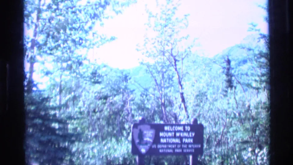 1977 Alaska 8mm frame showing the Mount McKinley National Park entrance sign amid trees with distant hills and bright sky.