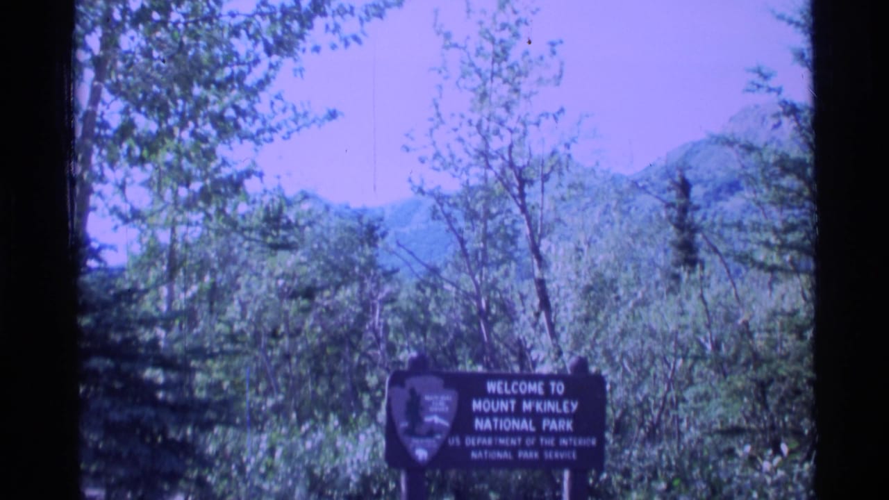 Park entrance sign with National Park Service shield, trees, hills, and blue-purple sky.