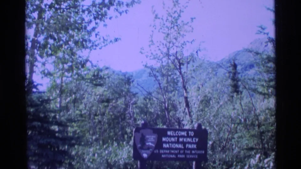 1977 Alaska 8mm frame with the Mount McKinley National Park sign low in frame, surrounded by trees and distant hills.