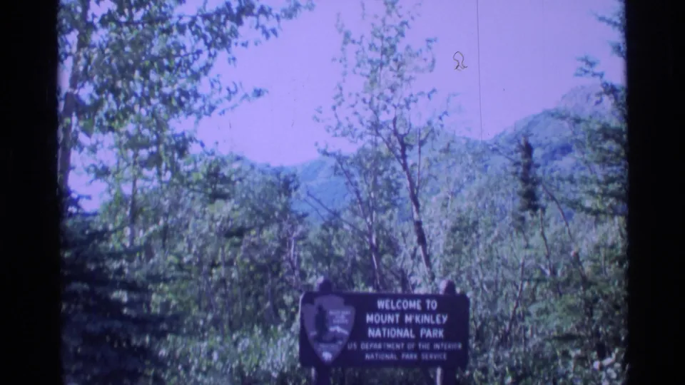1977 Alaska home-movie frame showing the Mount McKinley National Park entrance sign against trees, hills, and a color-shifted sky.
