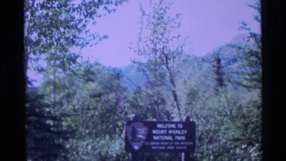 1977 Alaska home-movie frame showing a Mount McKinley National Park welcome sign amid trees with mountains in the background.
