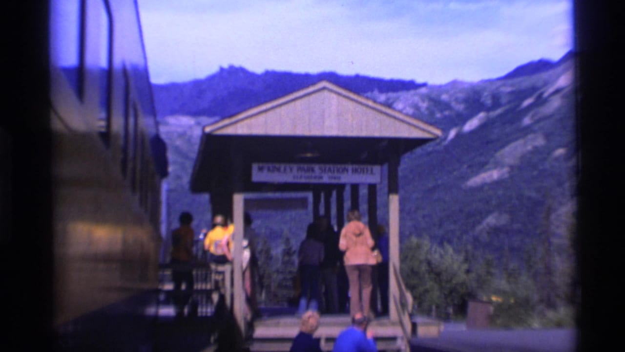 Passengers gather at the McKinley Park Station Hotel shelter in Alaska, with a dark train or bus side and mountains behind them.