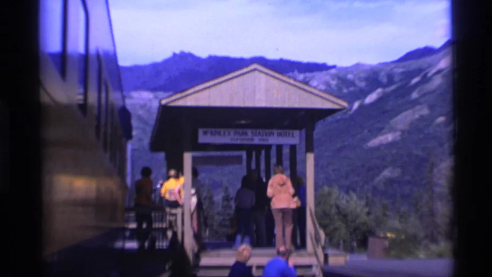 Passengers gather at the McKinley Park Station Hotel shelter in Alaska, with a dark train or bus side and mountains behind them.