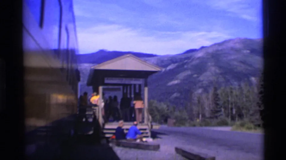 Alaska station stop with passengers near steps, a dark train or bus side at left, a paved road, trees, and mountains in the distance.