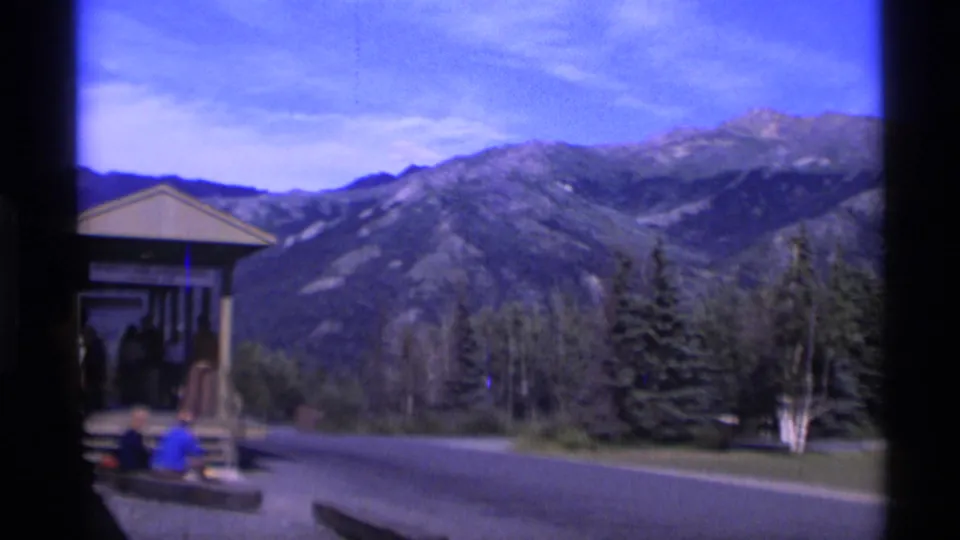 Mountain view at the Alaska station stop, with part of the shelter, passengers near the road, trees, and a dark vertical film edge.
