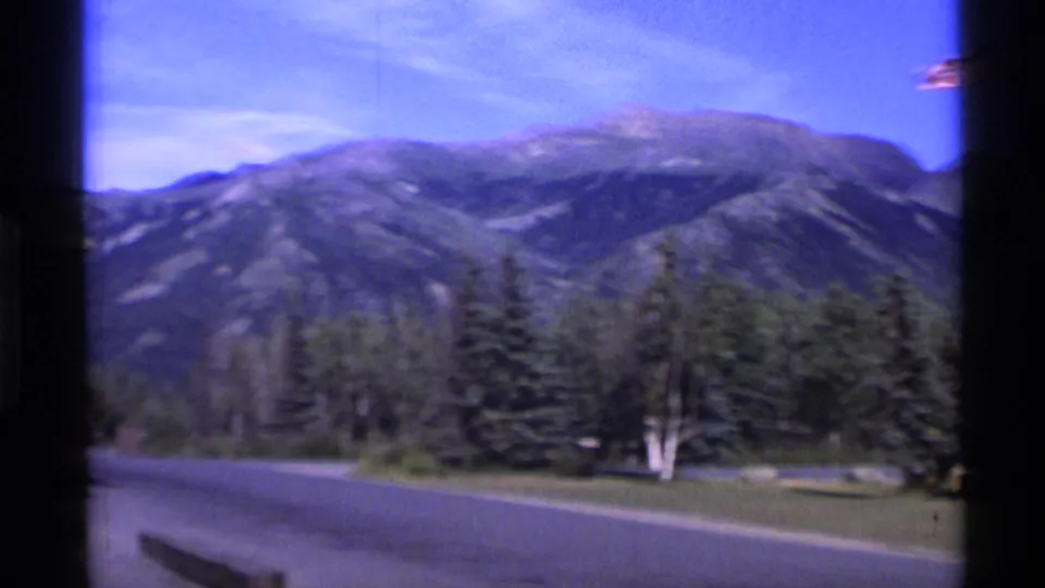 1977 Alaska 8mm frame showing mountains, forest, road edge, and dark gate borders from a home-movie travel shot.