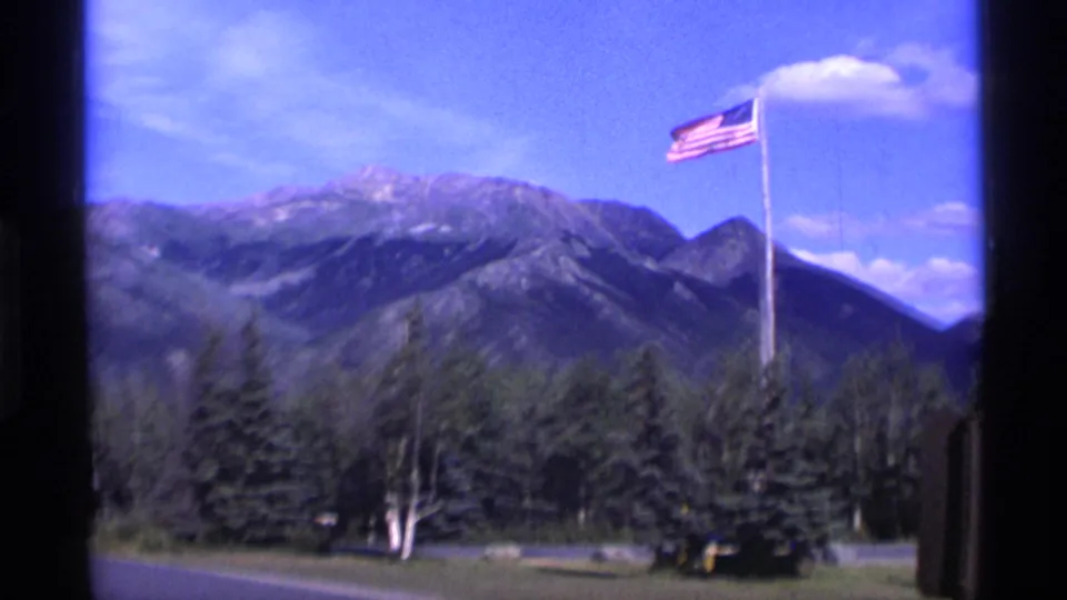 1977 Alaska travel frame with a U.S. flag flying near evergreen trees and mountain ridges under a blue sky.