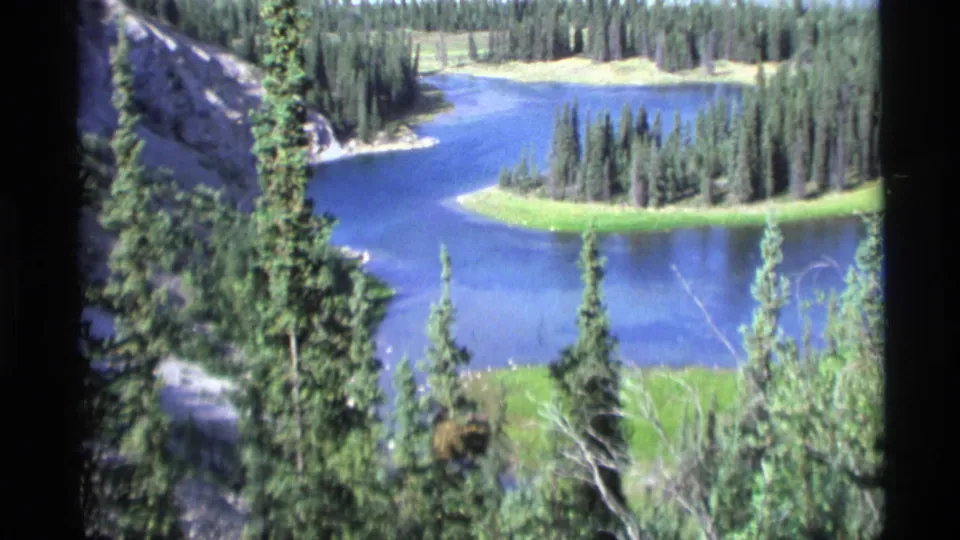 1977 Alaska 8mm frame showing a blue lake winding below an evergreen forest from an elevated overlook.