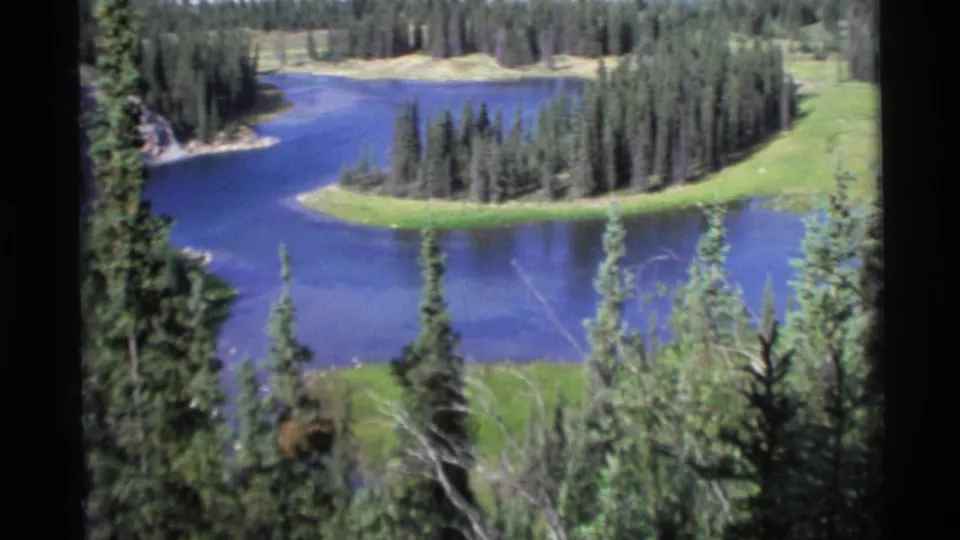 1977 Alaska home-movie view of a blue lake beside a tree-covered point and surrounding pine forest.