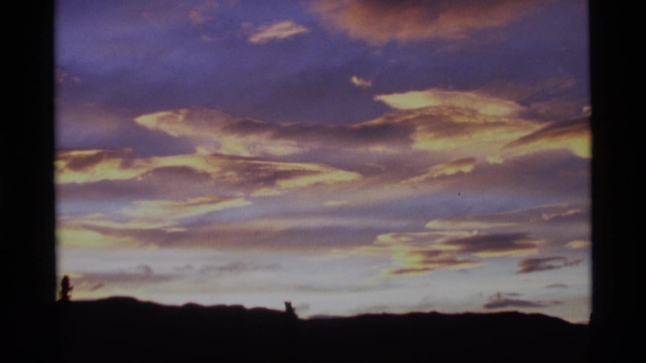 1977 Alaska home-movie still of luminous cloud layers above a low, dark ridge and scattered tree silhouettes.