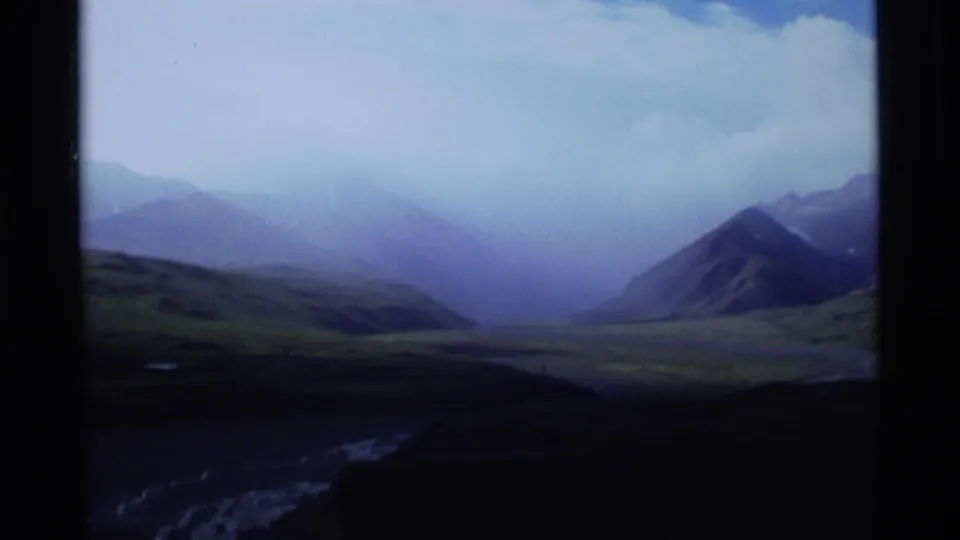 Alaska 8mm frame showing a distant mountain valley with mist or rain beneath a pale blue sky.