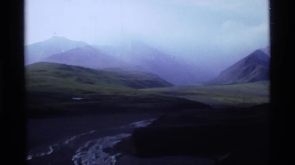 1977 Alaska 8mm frame showing mist-covered mountains, green hills, cloudy sky, and a river valley within dark projected-frame borders.