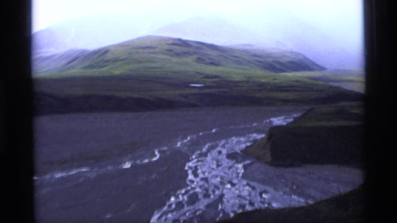 1977 Alaska home-movie frame with green slopes, a broad braided river channel, low cloud cover, and distant hazy mountains.