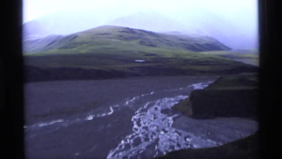 1977 Alaska home-movie frame with green slopes, a broad braided river channel, low cloud cover, and distant hazy mountains.
