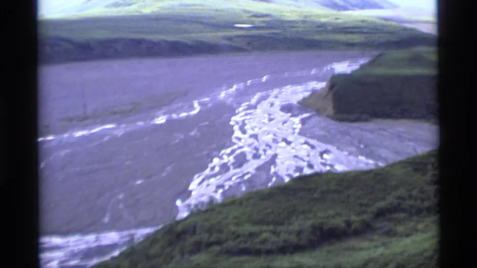 1977 Alaska 8mm frame looking over a pale braided river channel, green valley slopes, and overcast mountain terrain.