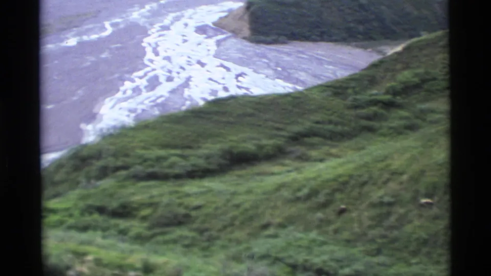 1977 Alaska home-movie frame with foreground grass, green hills, bright braided water channels, and hazy mountain landscape.