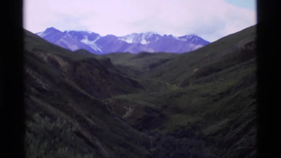 1977 Alaska 8mm mountain vista with snow-dusted peaks beyond dark green valley ridges.
