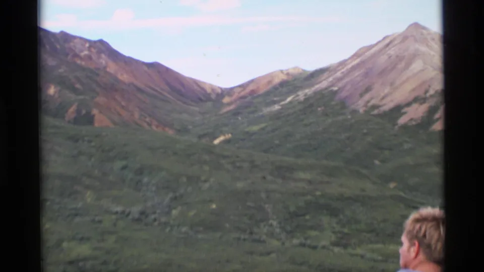 1977 Alaska 8mm frame showing a green mountain valley and reddish slopes, with a partial hiker's head at the lower edge.