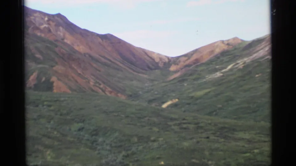 1977 Alaska 8mm view of a broad green valley and steep reddish-brown mountain slopes under a light blue sky.