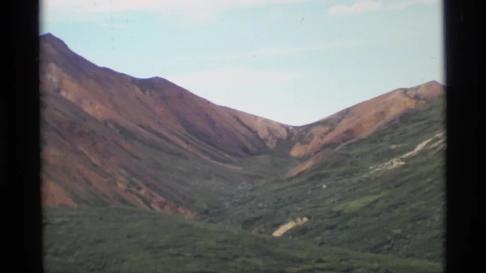 1977 Alaska home-movie frame of a mountain pass, with reddish slopes descending into green tundra-like ground.