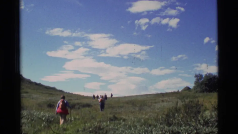 1977 Alaska 8mm frame showing hikers walking uphill across a grassy meadow under scattered clouds.