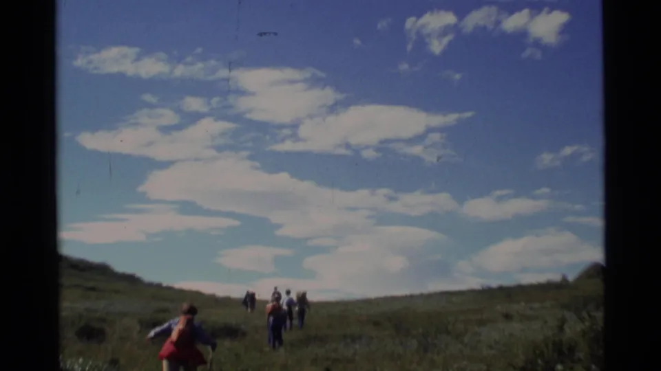 A distant hiker with a red pack trails a small group crossing an Alaskan meadow in 1977 home-movie footage.
