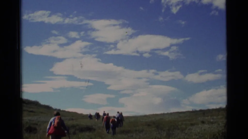 1977 Alaska 8mm frame of a hiking group moving over a grassy slope with a wide cloud-filled sky above.