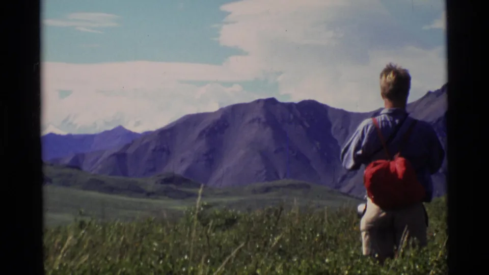 A hiker with a red backpack stands in an Alaskan meadow facing mountain ridges in 1977 8mm footage.