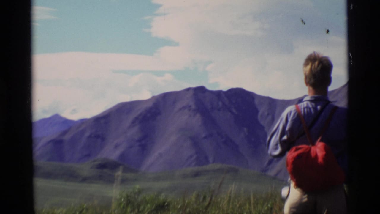 1977 Alaska 8mm frame of a hiker seen from behind with a red backpack, looking toward broad mountains under clouds.